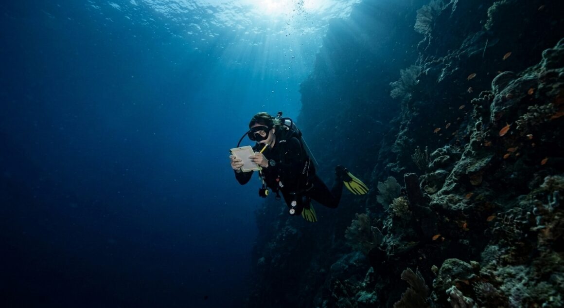 Scuba diver checking dive computer at depth along a deep blue drop-off wall